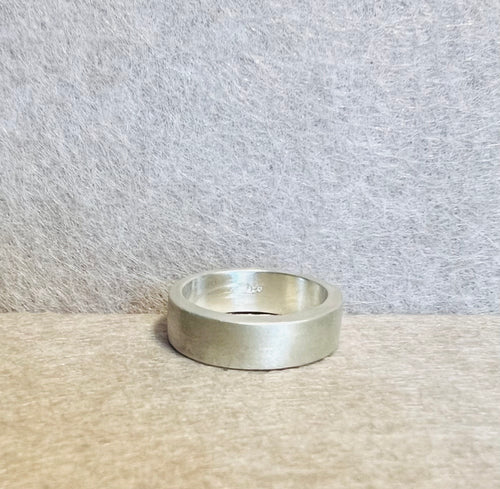 Wide silver-colored band ring with a brushed surface, photographed on beige fabric against a textured gray background.