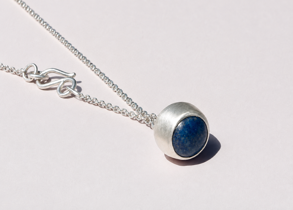 Blue-stone pendant in a rounded silver-colored setting, with chain and hook clasp arranged diagonally on a warm ivory background.
