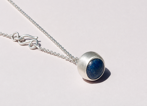 Blue-stone pendant in a rounded silver-colored setting, with chain and hook clasp arranged diagonally on a warm ivory background.