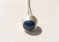 Rounded pendant with a deep blue stone and gold-colored flecks in a textured silver-colored setting, photographed on warm ivory.