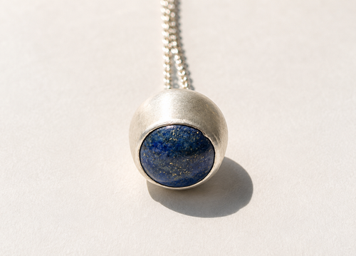 Rounded pendant with a deep blue stone and gold-colored flecks in a textured silver-colored setting, photographed on warm ivory.