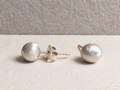 Pair of rounded, textured silver-colored stud earrings with posts and a butterfly back, photographed on an ivory fabric surface.