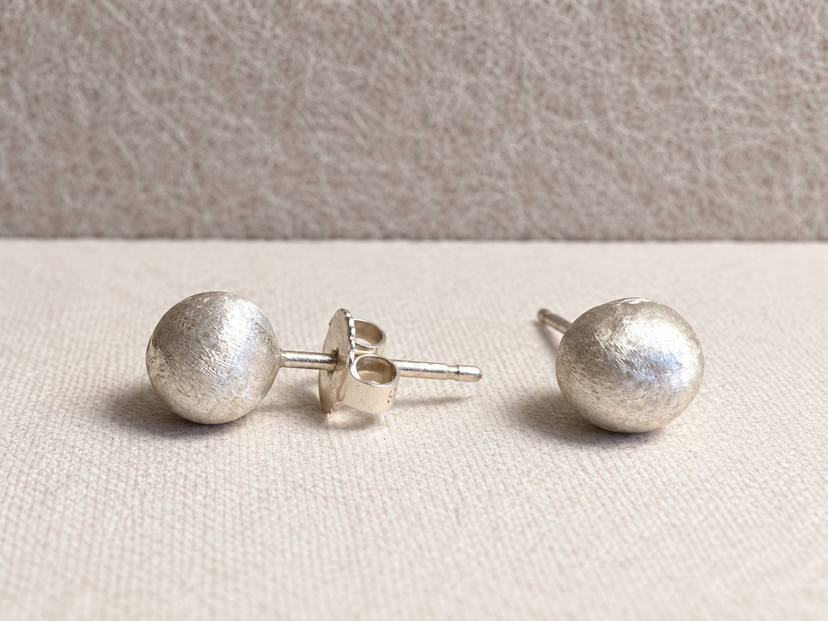 Pair of rounded, textured silver-colored stud earrings with posts and a butterfly back, photographed on an ivory fabric surface.